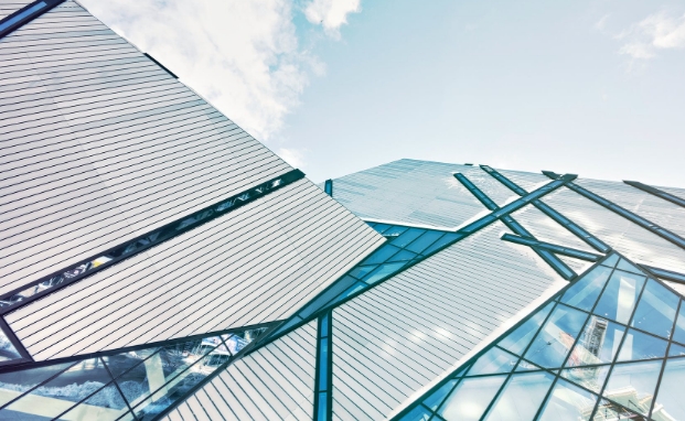 Estructura arquitectónica moderna con paneles de vidrio y metal dispuestos en ángulos agudos y líneas diagonales, creando un diseño geométrico abstracto. El cielo azul con algunas nubes es visible en el fondo.